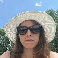 Une personne portant des lunettes de soleil et un chapeau à large bord est à l'extérieur. Il y a des arbres et un ciel bleu avec quelques nuages en arrière-plan.