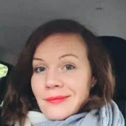 Femme aux cheveux bruns mi-longs et au rouge à lèvres rouge, portant un foulard, assise dans une voiture.